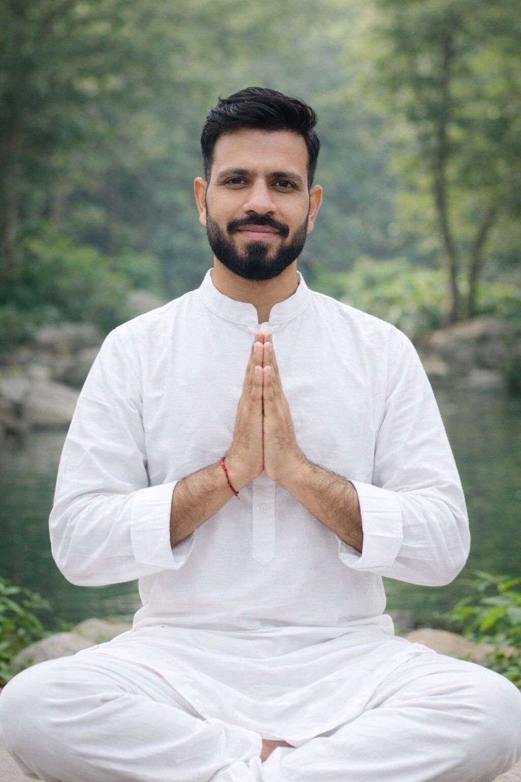 Dayaram seated in meditation with hands in namaste, dressed in white by a forest river
